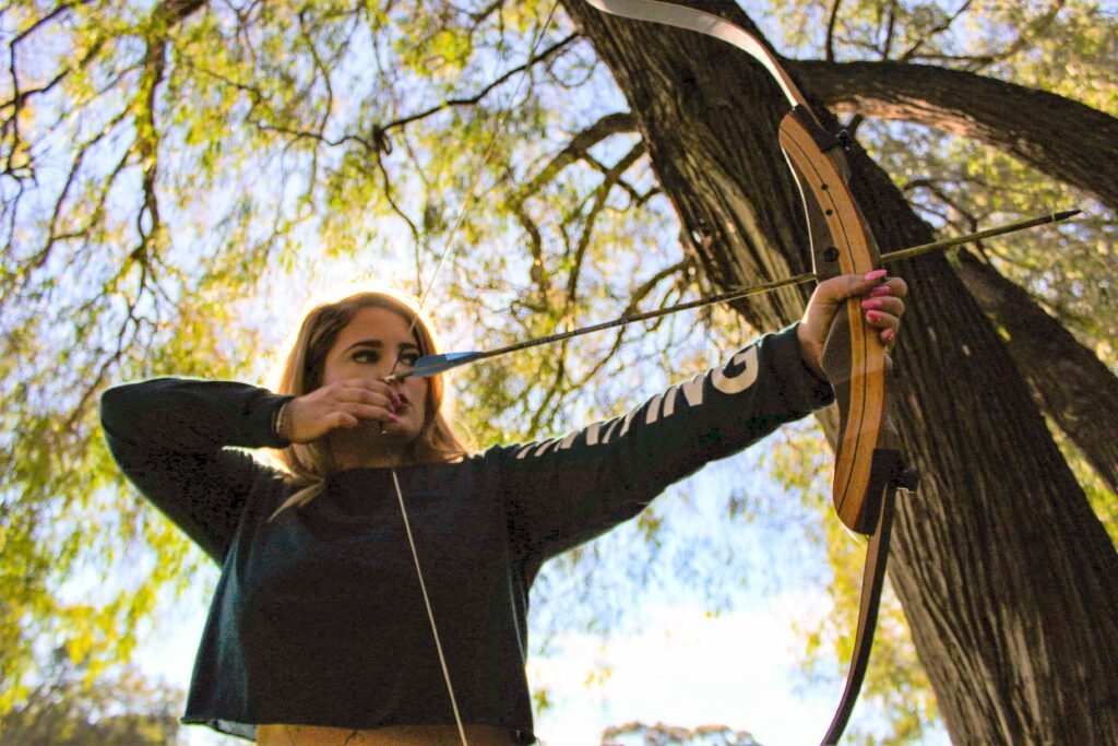 Right handed Archer with her bow drawn and arrow pointing downrange. The photo is set outside with trees in the background at a low angle.